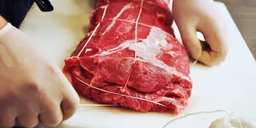Mains gantées ficelant une pièce de viande pour en maintenir la forme